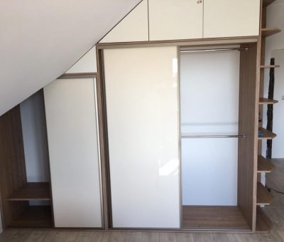 Nowoczesna zabudowa wnęki z drzwiami przesuwnymi, idealna do wykorzystania przestrzeni pod skośnym sufitem. Szafa ma dwie główne sekcje z drzwiami przesuwnymi. Jedna z nich jest otwarta, ukazując drążek na ubrania oraz półki po prawej stronie. Dodatkowe komory przechowawcze znajdują się powyżej głównych sekcji. Całość wykonana z drewna o jasnym wykończeniu, co nadaje wnętrzu nowoczesny i funkcjonalny charakter.