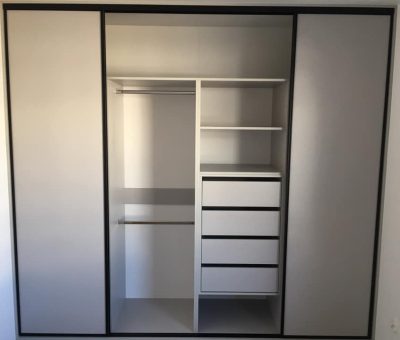 Nowoczesna zabudowa wnęki z trzema sekcjami wyposażonymi w drzwi przesuwne. Lewa sekcja posiada drążek na ubrania, środkowa łączy półki i szuflady z dodatkowym drążkiem, natomiast prawa sekcja pozostaje zamknięta. Meble są utrzymane w minimalistycznym, monochromatycznym stylu, z czystymi liniami i kolorystyką w odcieniach bieli i czerni. Idealne rozwiązanie dla osób ceniących nowoczesny design i funkcjonalność.